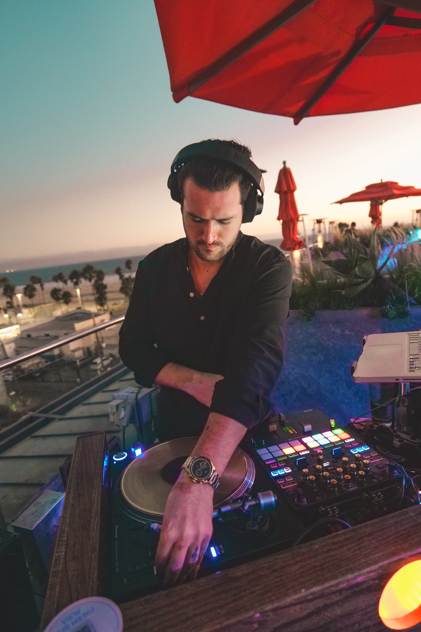 Miles at the turntables on a rooftop at dusk, with the ocean and palm trees behind him.