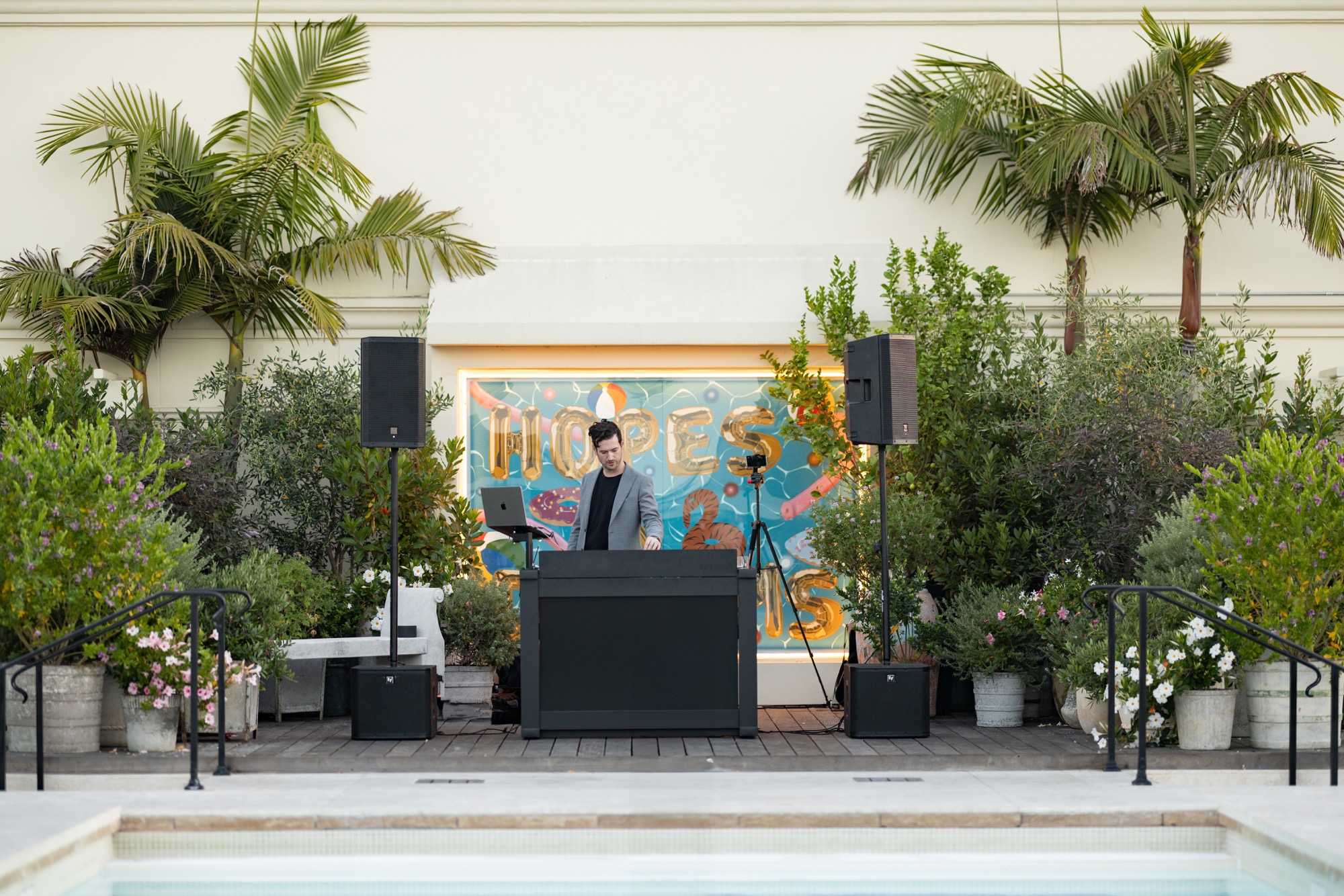 A DJ at a booth beside a hotel pool before any guests arrive, speakers on stands either side, potted plants and a painted backdrop behind.