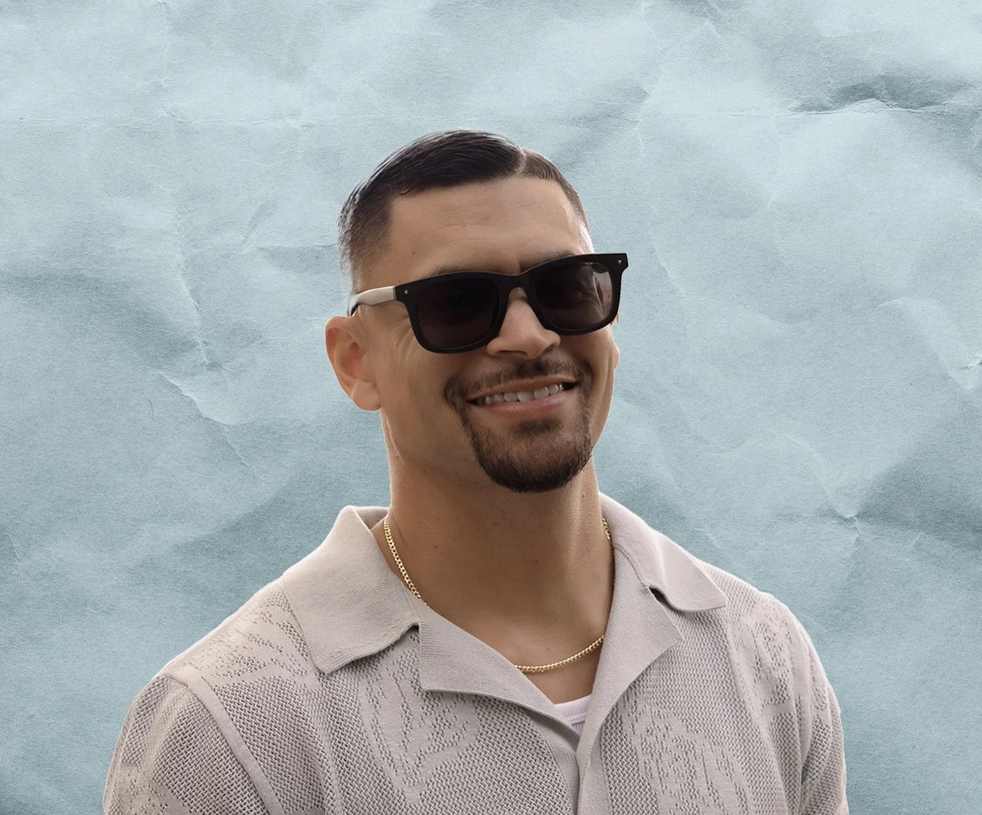 Brandon Cruz in sunglasses and an open knit shirt, smiling, against a pale paper backdrop.