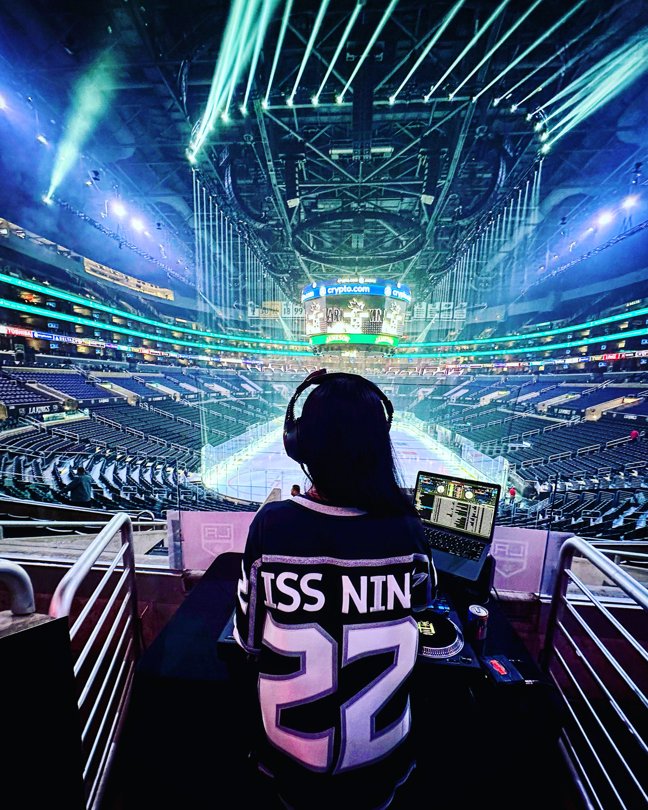 DJ Miss Ninja from behind in an LA Kings jersey with 22 on the back, at the decks high in the arena, lights sweeping the empty rink below.