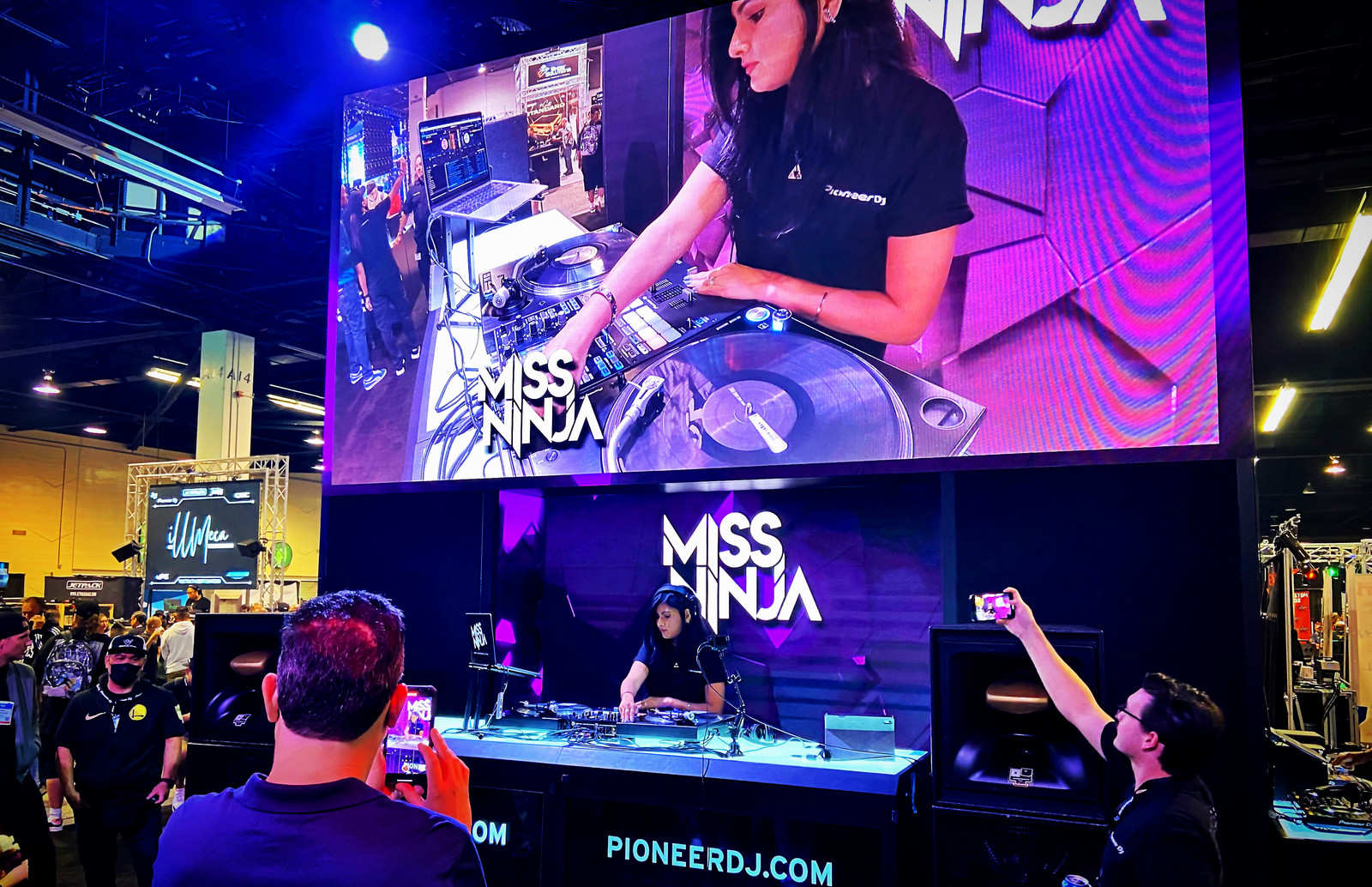 DJ Miss Ninja playing the Pioneer DJ booth, a giant screen of her hands on the decks above, the crowd filming on their phones.