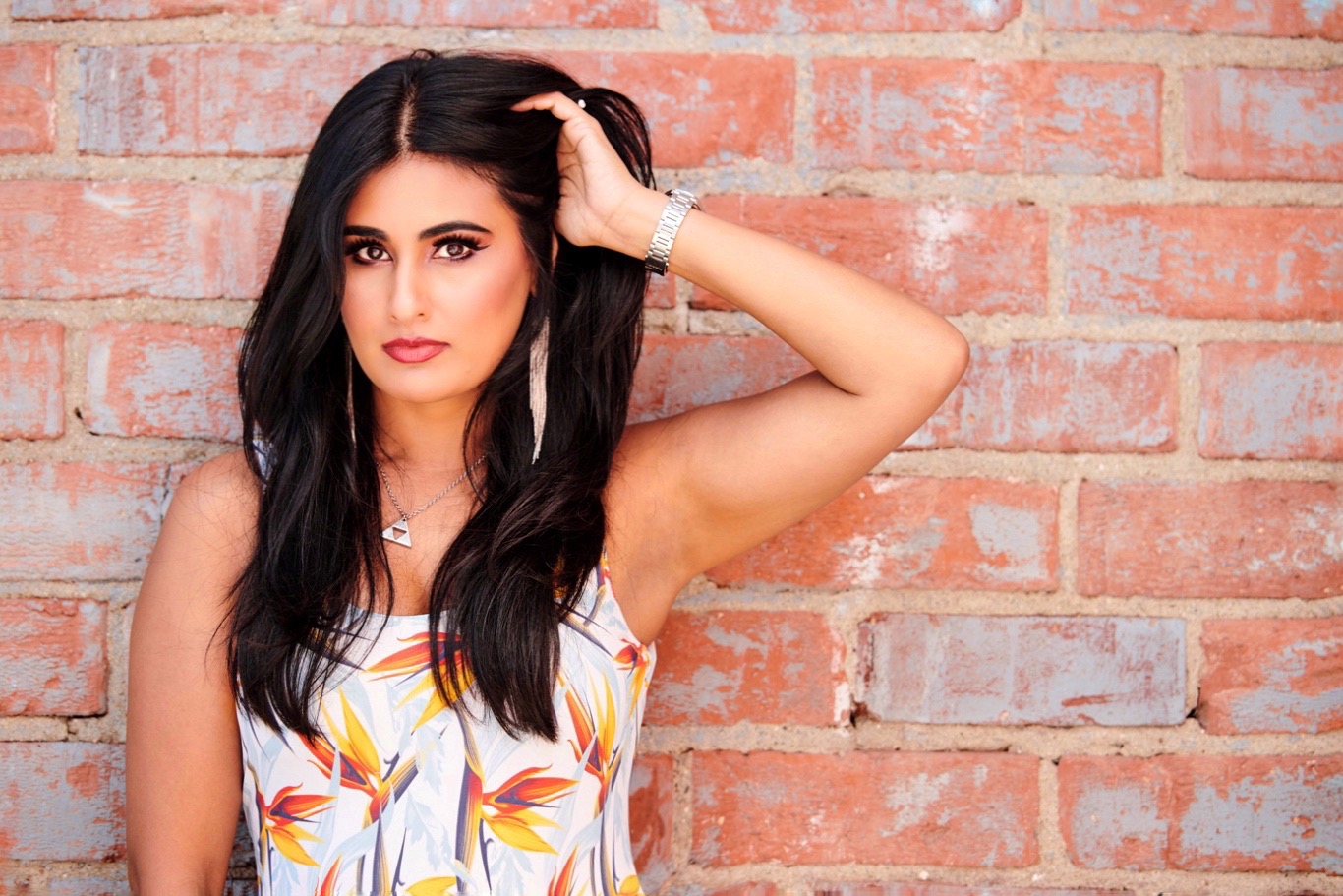 DJ Miss Ninja against a red brick wall, one hand in her long dark hair, looking straight at the camera.