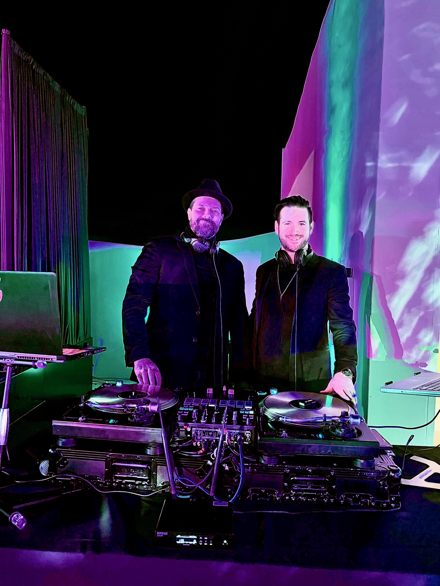 Lee Dyson in a black hat and Miles side by side behind two turntables and a mixer, both smiling, under green and violet light.