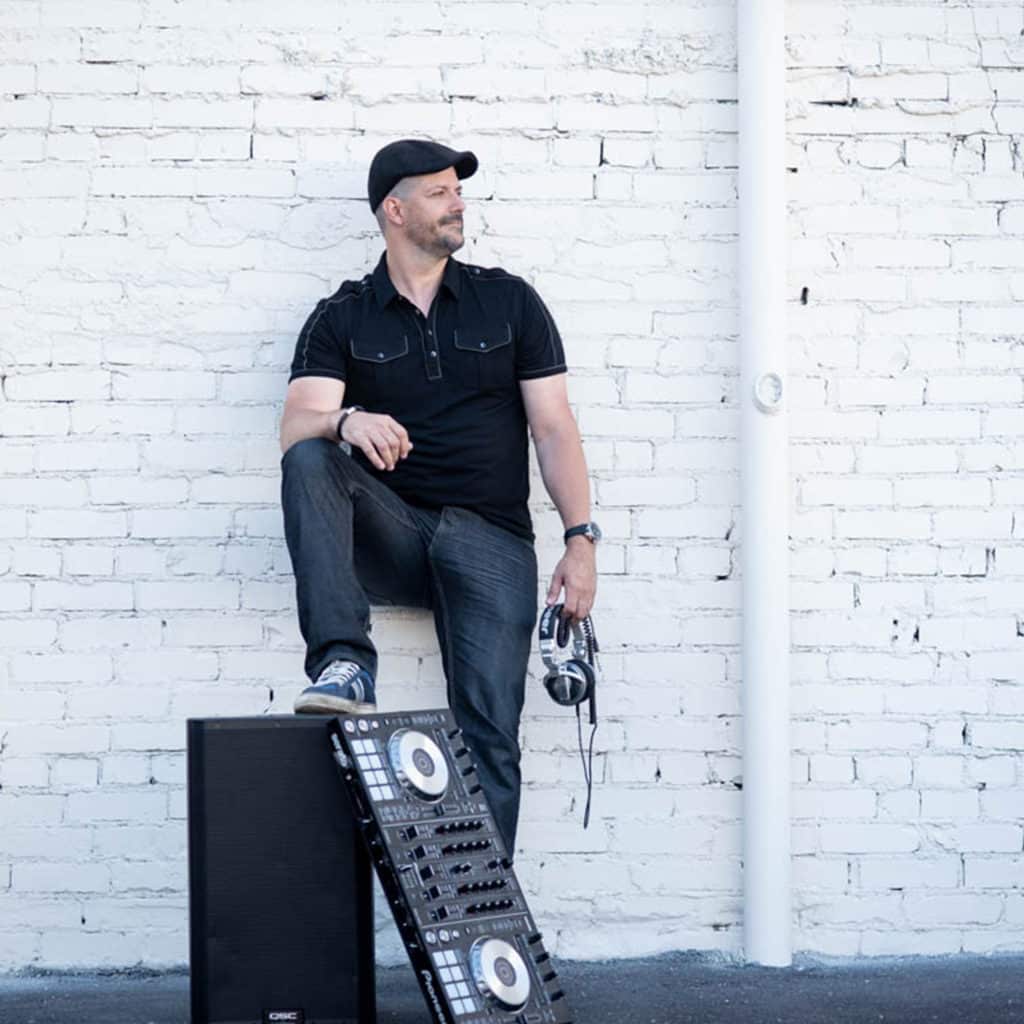 Lee Dyson in a flat cap and black shirt, headphones in hand, seated on a speaker against a white brick wall.