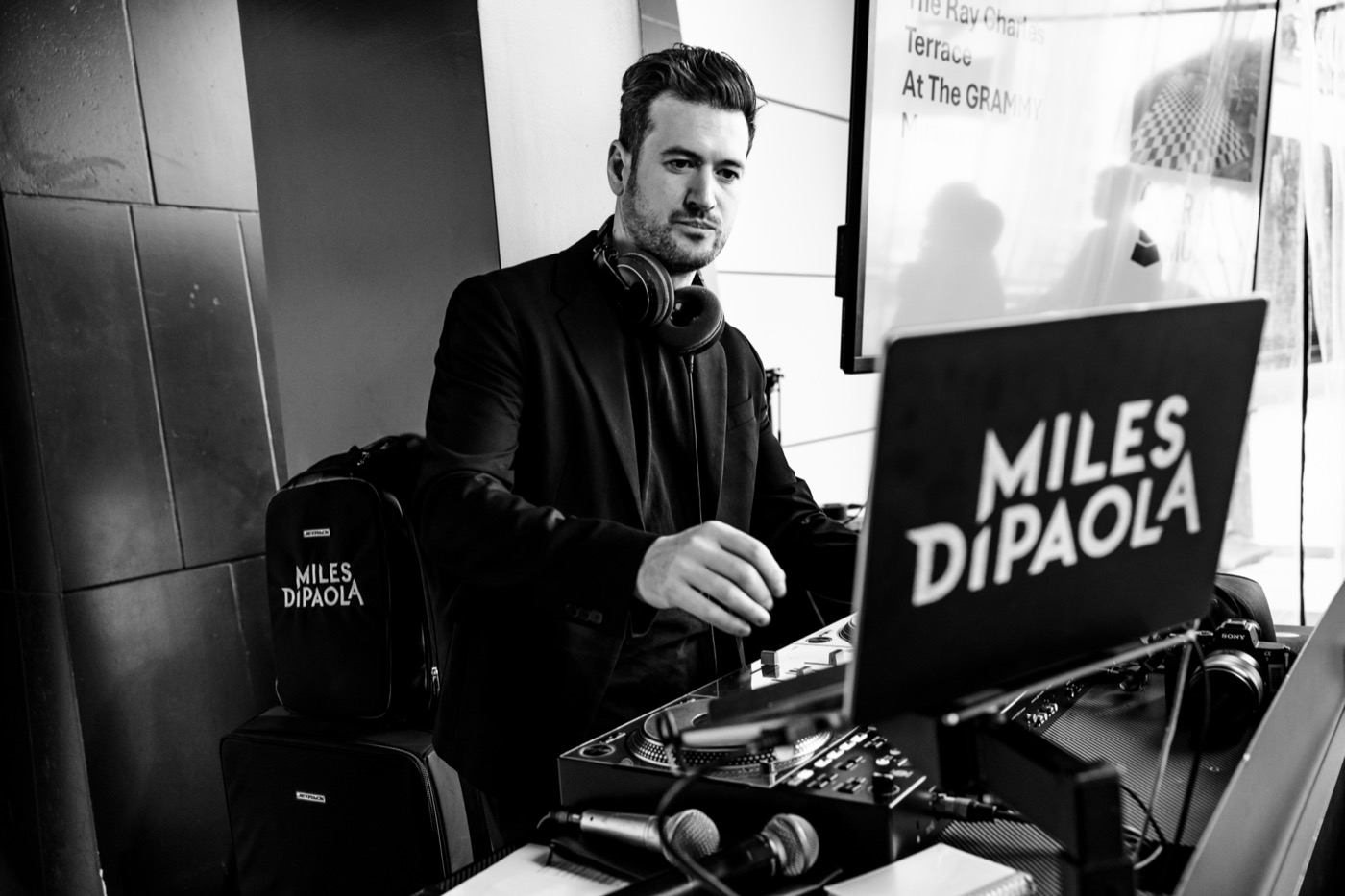 Miles at the decks in black and white, headphones around his neck and one hand on the controls.