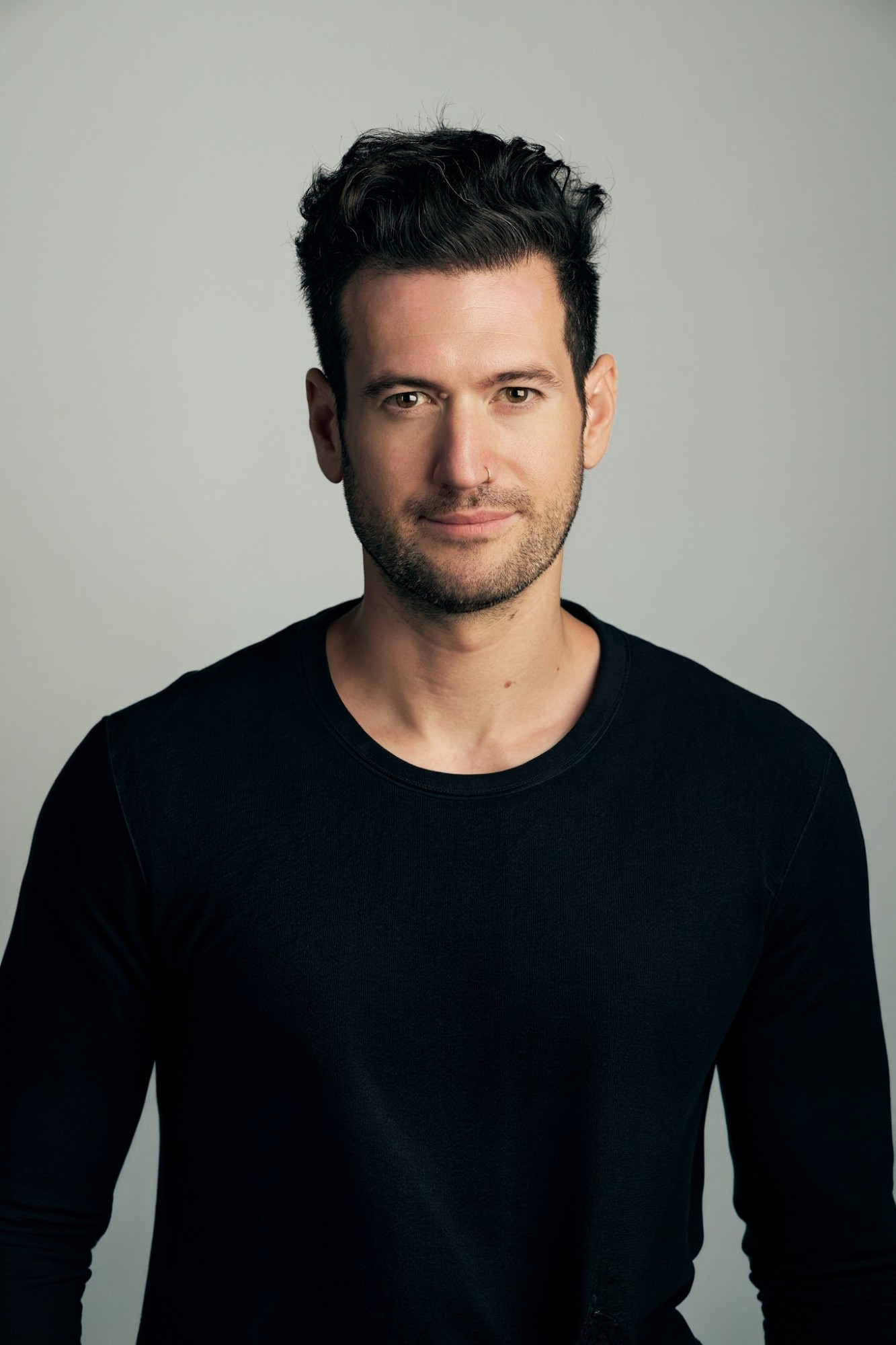 Miles DiPaola in a black long-sleeve shirt against a pale grey studio backdrop, looking at the camera.