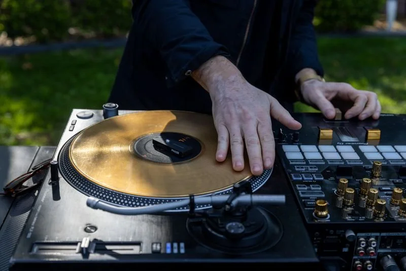 A hand on a gold record on a turntable, outdoors in daylight, a mixer beside it.