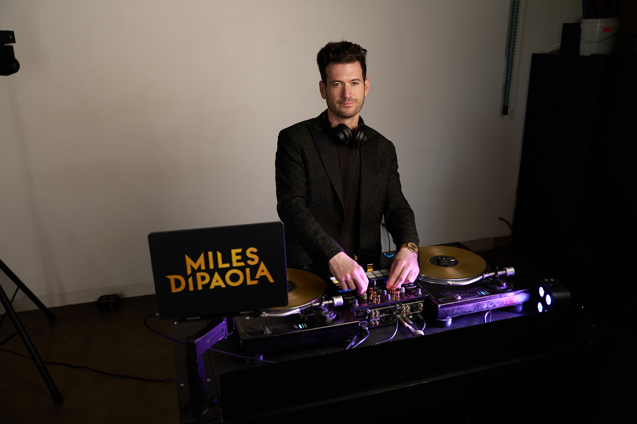 Miles behind two turntables with gold records on them and a mixer between, headphones around his neck, a lit sign with his name beside the decks.