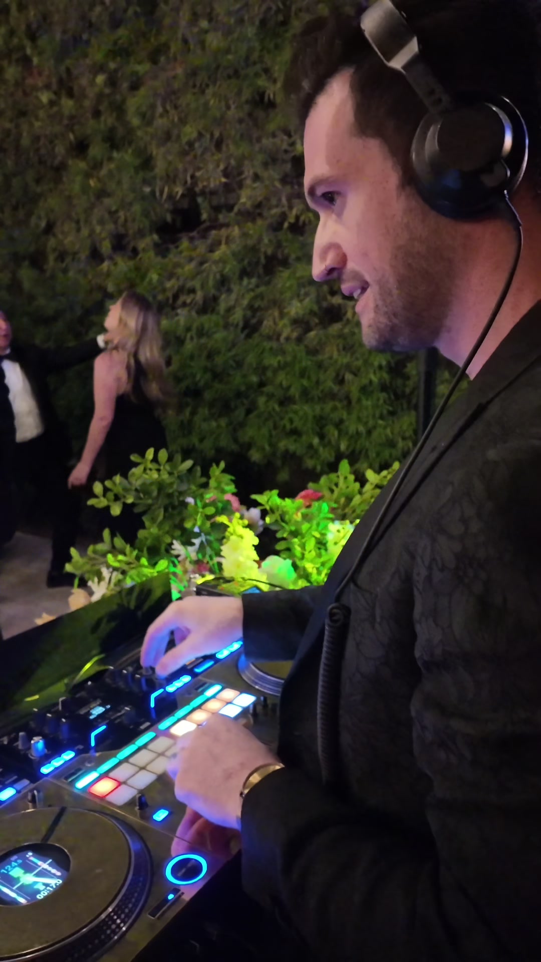 Over the DJ’s shoulder: headphones on, hands on the lit controller, guests and a hedge beyond the booth.