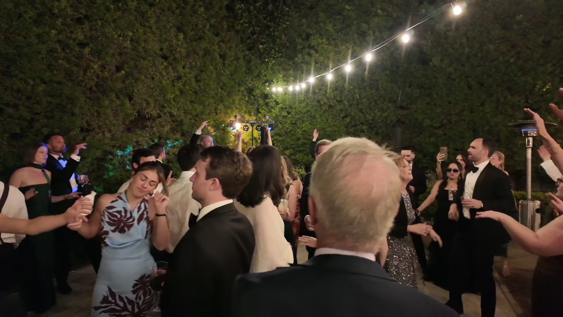 A full dance floor under strings of warm lights in a garden at night, guests in black tie facing the booth.