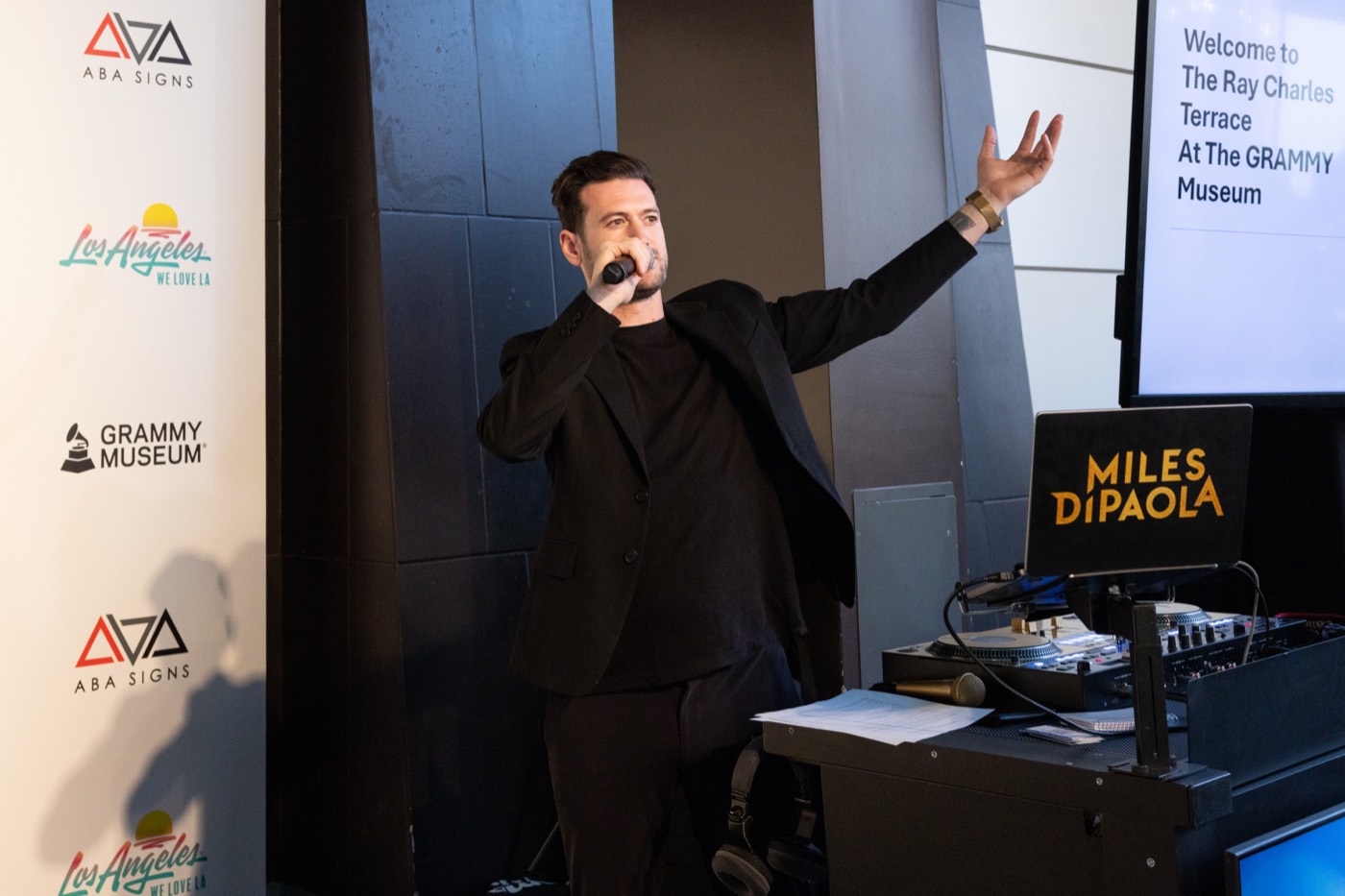 Miles speaking into a handheld microphone with one arm open, beside the decks and an event backdrop.
