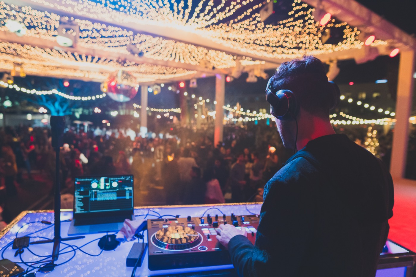 A DJ at the decks, seen from behind, looking out over a full floor under warm strings of lights.