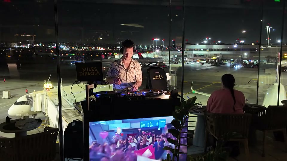 A DJ booth in front of tall windows at night, with aircraft and apron lights outside.
